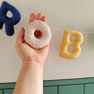 May include: A garland of soft, plush fabric letters strung on a white cord. The letters spell out the word ROBE, featuring a dark blue R, a cream-coloured O, a yellow B, and a light green E, all with a textured, fuzzy surface finish.