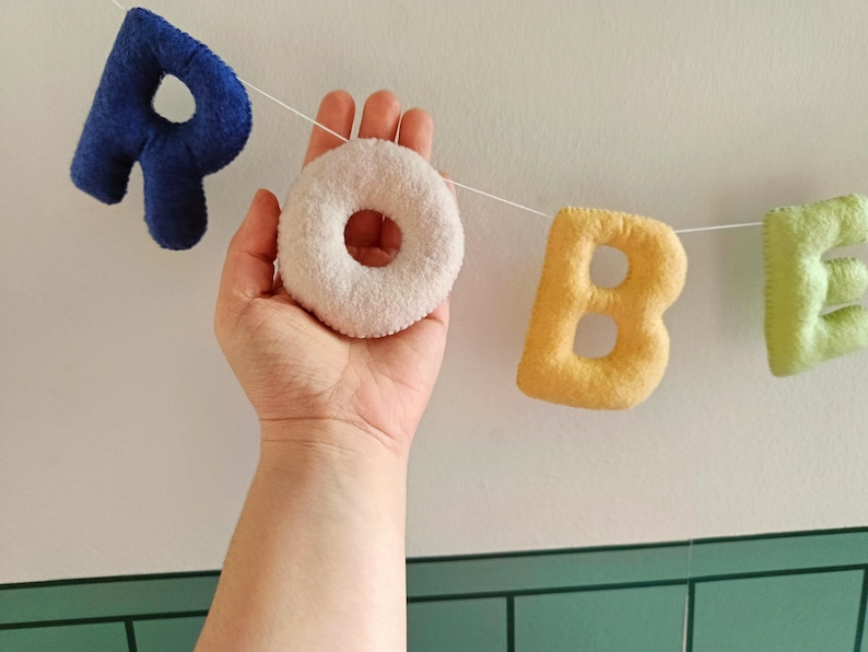 May include: A garland of soft, plush fabric letters strung on a white cord. The letters spell out the word ROBE, featuring a dark blue R, a cream-coloured O, a yellow B, and a light green E, all with a textured, fuzzy surface finish.