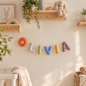 May include: A decorative wall garland featuring plush, textured fabric letters spelling the name OLIVIA in orange, light grey, green, blue, yellow, and purple. Each letter is suspended on a thin white string, creating a colourful banner display.
