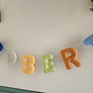 May include: A decorative garland featuring plush, soft-textured fabric letters spelling out the name ROBERT. Each letter is a different colour, including dark blue, white, yellow, light green, orange, and light blue, all strung on a thin white cord.