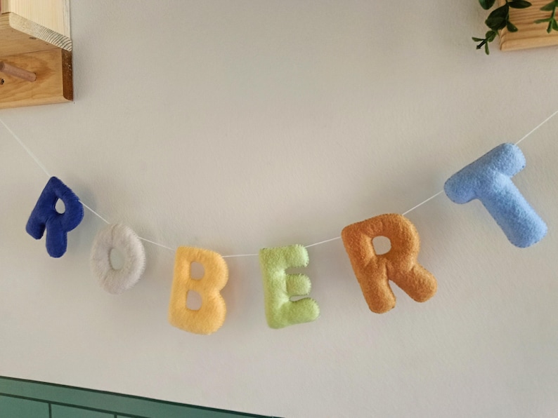 May include: A decorative garland featuring plush, soft-textured fabric letters spelling out the name ROBERT. Each letter is a different colour, including dark blue, white, yellow, light green, orange, and light blue, all strung on a thin white cord.