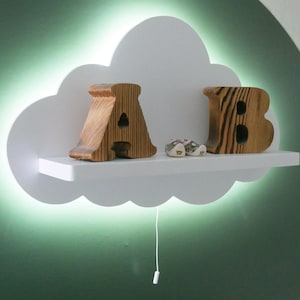 Pode incluir: Luz noturna branca em forma de nuvem com uma prateleira embutida. A prateleira tem duas letras de madeira, "A" e "B", e um par de sapatos pequenos. A luz está acesa e projeta um brilho verde.
