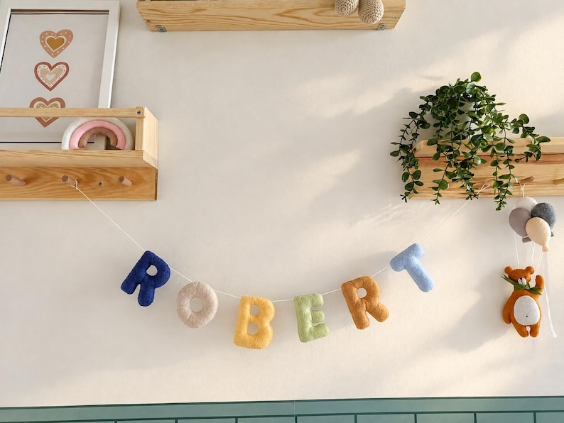 May include: A decorative garland featuring plush, fabric-covered letters spelling the name ROBERT in various colours including dark blue, beige, yellow, light green, brown, and light blue, suspended on a white string with a small bear and balloons.