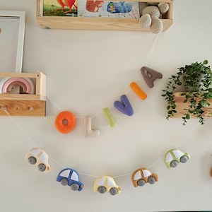 May include: A decorative garland featuring felt letters spelling the name OLIVIA in orange, beige, green, purple, and brown, suspended above a second garland of felt cars in various colours, with a small brown bear and balloons at the end.