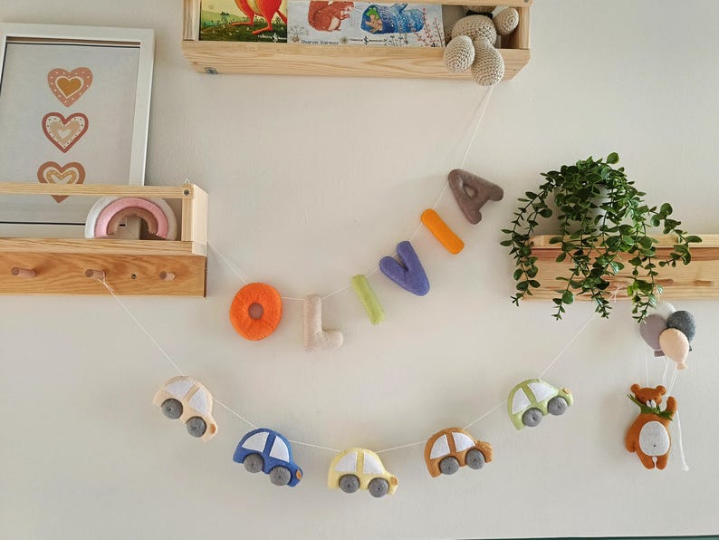 May include: A decorative garland featuring felt letters spelling the name OLIVIA in orange, beige, green, purple, and brown, suspended above a second garland of felt cars in various colours, with a small brown bear and balloons at the end.