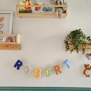 May include: A felt letter garland spelling the name ROBERT in various colours, including blue, beige, yellow, green, orange, and light blue. The letters are strung on a white cord, accompanied by a small felt bear and balloon ornament.