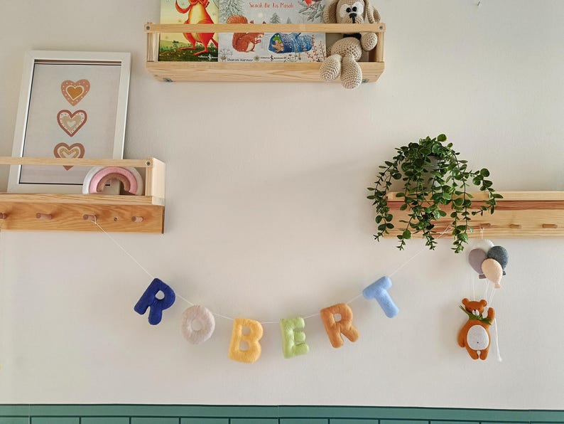 May include: A felt letter garland spelling the name ROBERT in various colours, including blue, beige, yellow, green, orange, and light blue. The letters are strung on a white cord, accompanied by a small felt bear and balloon ornament.