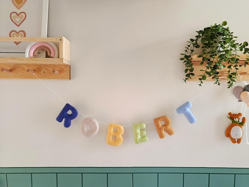 May include: A decorative garland featuring individual plush fabric letters spelling the name ROBERT, each in a different colour including dark blue, cream, yellow, light green, orange, and light blue, all strung together on a thin white cord.