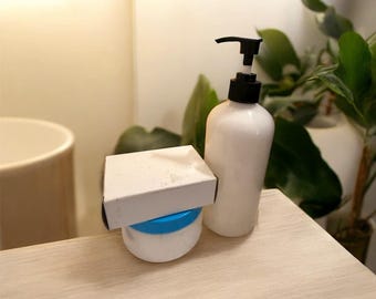 May include: A white lotion bottle with a black pump, a small white box, and a white jar with a blue lid sit on a light-colored wooden surface. Green plants are in the background.