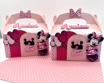 May include: Two pink party favor boxes with handles, decorated with Minnie Mouse and the name "Azalea". Each box features a pink bow, a number "2", and a Minnie Mouse figure. The boxes sit on a pink polka dot surface.