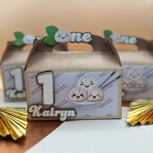 May include: Brown cardboard favor boxes with a dumpling theme. Each box has a number 1, the name "Kairyn", and a dumpling illustration. The handle has the word "one" and a dumpling graphic. Gold paper fans are in the foreground.