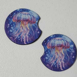 May include: Two round car coasters with a watercolor jellyfish design. The jellyfish is depicted in shades of pink, purple, and yellow, set against a dark blue background with white speckles. Each coaster has a semi-circular cutout.