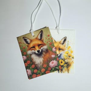May include: Two rectangular tags with watercolor illustrations of foxes. One fox is surrounded by pink and red flowers, the other by yellow flowers. The tags are connected by a white string.