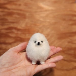 Op de afbeelding: Een kleine, pluizige witte Pomeriaan hond figuur met zwarte ogen en neus. De handgemaakte vilten hond staat op een open handpalm, tegen een vage houten achtergrond.