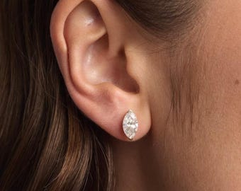 May include: A marquise-cut diamond stud earring set in a gold bezel. The earring is shown on an ear, highlighting its shape and sparkle. The setting appears to be a warm yellow gold.