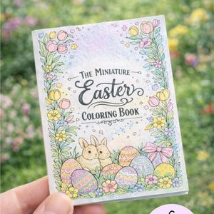 May include: A miniature Easter coloring book held in a hand. The cover features the text "The Miniature Easter Coloring Book" with illustrations of bunnies, Easter eggs, and flowers. The book has 6 coloring pages.