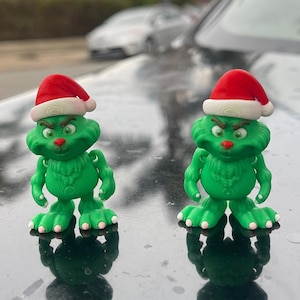 May include: Two green Grinch figurines wearing red Santa hats are standing on a black car. The Grinch figures have a grumpy expression on their faces.