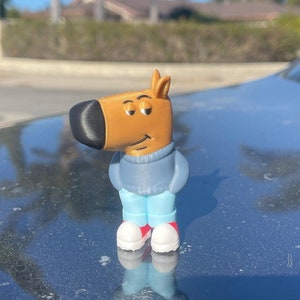 May include: A small, tan figurine of a cartoon dog with a black nose, wearing a blue sweater, light blue pants, and white shoes with red accents. The figurine is standing on a dark surface, with a blurred background of trees and a blue sky.