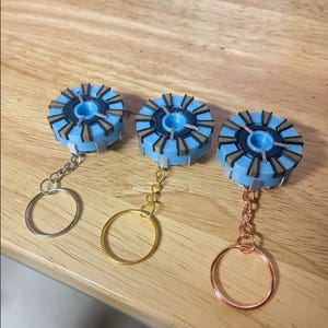 May include: Three blue and gold keychains with different colored key rings. The keychains are shaped like a circular disc with a central hole and a pattern of triangles around the edge.