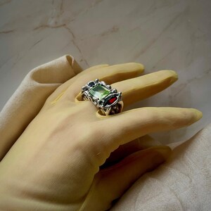 May include: A silver ring with a rectangular green gemstone and a smaller red gemstone. The ring has an ornate, textured design around the stones. The ring is displayed on a gloved hand.