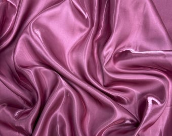 May include: Close-up of a flowing, lustrous fabric in a deep rose color. The material has a silky sheen, with soft folds and highlights that create a sense of depth and movement. The fabric appears to be a satin or similar material, suitable for apparel or decorative use.