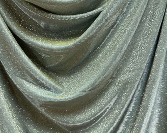 May include: Close-up of shimmering silver fabric with a textured, ribbed pattern. The fabric drapes in soft folds, catching the light and creating a sparkling effect. A thin gold stripe runs along the top edge.