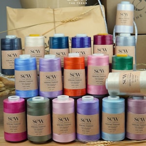 May include: A collection of Sew The Trend polyester sewing thread spools in various colors including blue, pink, orange, green, purple, and grey. Each spool features a tan paper label with the text Sew The Trend Sewing Thread 6000 Yards.