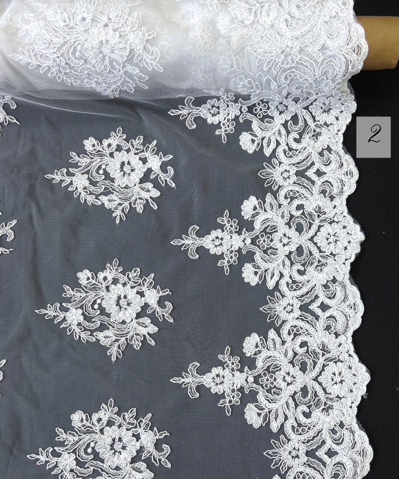 May include: White floral lace fabric with a scalloped edge. The lace is delicate and intricate, with a repeating pattern of flowers and leaves.
