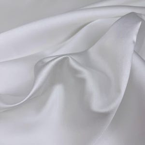 May include: White satin fabric with a smooth, silky texture. The fabric is draped and folded, creating soft curves and folds.