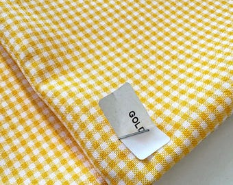 May include: Close-up of yellow and white gingham fabric. The fabric is folded, with a tag that reads "GOLD" attached. The pattern is a classic checkered design, perfect for various crafting projects or home decor.