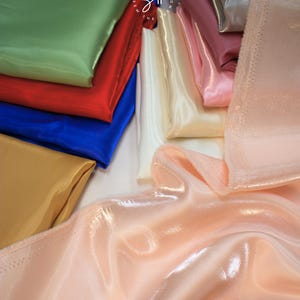 May include: A collection of folded satin fabric in various colors, including green, red, blue, gold, white, pink, and peach. The fabric has a shiny, smooth texture, and the folds create shadows and highlights.
