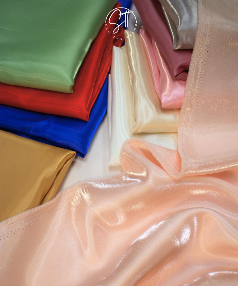 May include: A collection of folded satin fabric in various colors, including green, red, blue, gold, white, pink, and peach. The fabric has a shiny, smooth texture, and the folds create shadows and highlights.