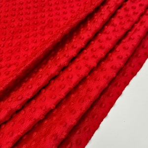 May include: Close-up of a folded red minky fabric. The fabric has a soft, plush texture with raised dot pattern. The folds create shadows, highlighting the texture and depth of the material. The bright red color is consistent throughout the fabric.