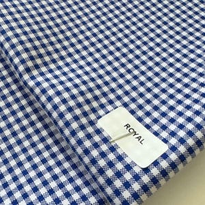 May include: Close-up of blue and white gingham fabric. The fabric is folded, showing the pattern's repeating squares. A white tag with the word "ROYAL" is attached to the fabric.