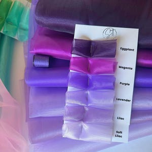 May include: A color swatch card featuring six sheer organza fabric samples in shades of eggplant, magenta, purple, lavender, lilac, and soft lilac. Each fabric piece is pinned to a white card with corresponding text labels. A spool of purple thread is visible.