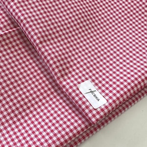 May include: A folded piece of fabric with a red and white gingham pattern. The fabric has a small, square check design. A white tag with the word "FUCSHIA" is attached to the fabric.