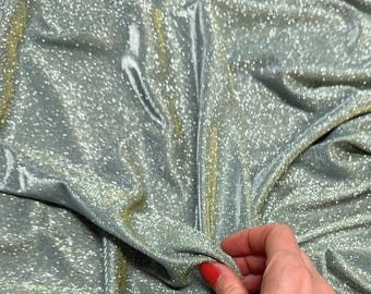 May include: Close-up of shimmering fabric in shades of silver and gold. The material has a textured, reflective surface with a subtle sheen. The fabric is draped, creating soft folds and shadows. The overall effect is luxurious and eye-catching.