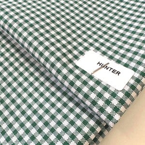 May include: Close-up of green and white gingham fabric. The fabric is folded, revealing a pattern of small, evenly spaced squares. A white tag with the word "HUNTER" is attached to the fabric.