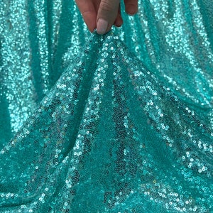 Puede incluir: Tela de lentejuelas turquesa con un acabado brillante. La tela está drapeada y doblada, mostrando la textura y el brillo de las lentejuelas.