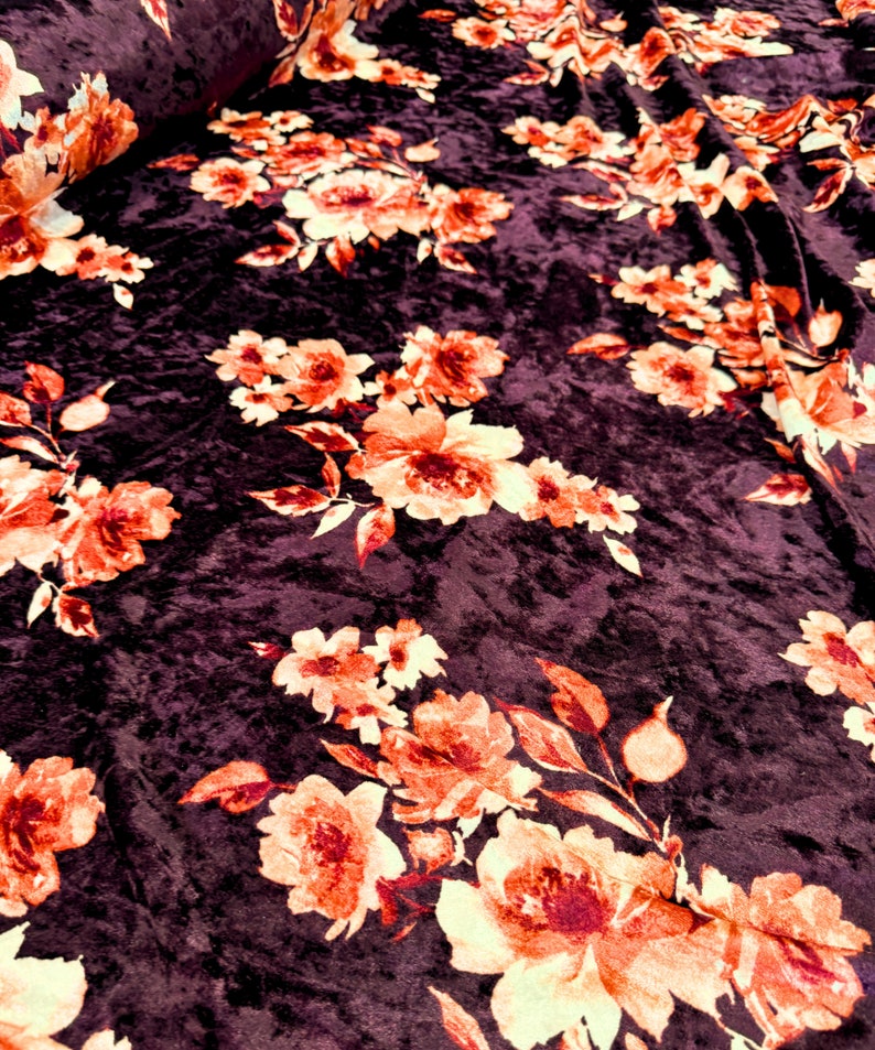 May include: Close-up of a dark purple velvet fabric with a floral pattern. The flowers are orange and yellow with a white outline. The fabric has a soft, plush texture.