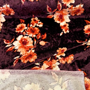 May include: A close-up of a burgundy velvet fabric with a floral pattern in shades of orange and cream. The fabric is folded in half, revealing a lighter-colored fabric underneath.