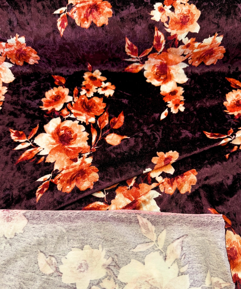 May include: A close-up of a burgundy velvet fabric with a floral pattern in shades of orange and cream. The fabric is folded in half, revealing a lighter-colored fabric underneath.