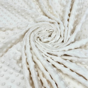 May include: Close-up of a white, soft, textured fabric with a raised dot pattern. The fabric is folded and twisted, showing the texture and softness.