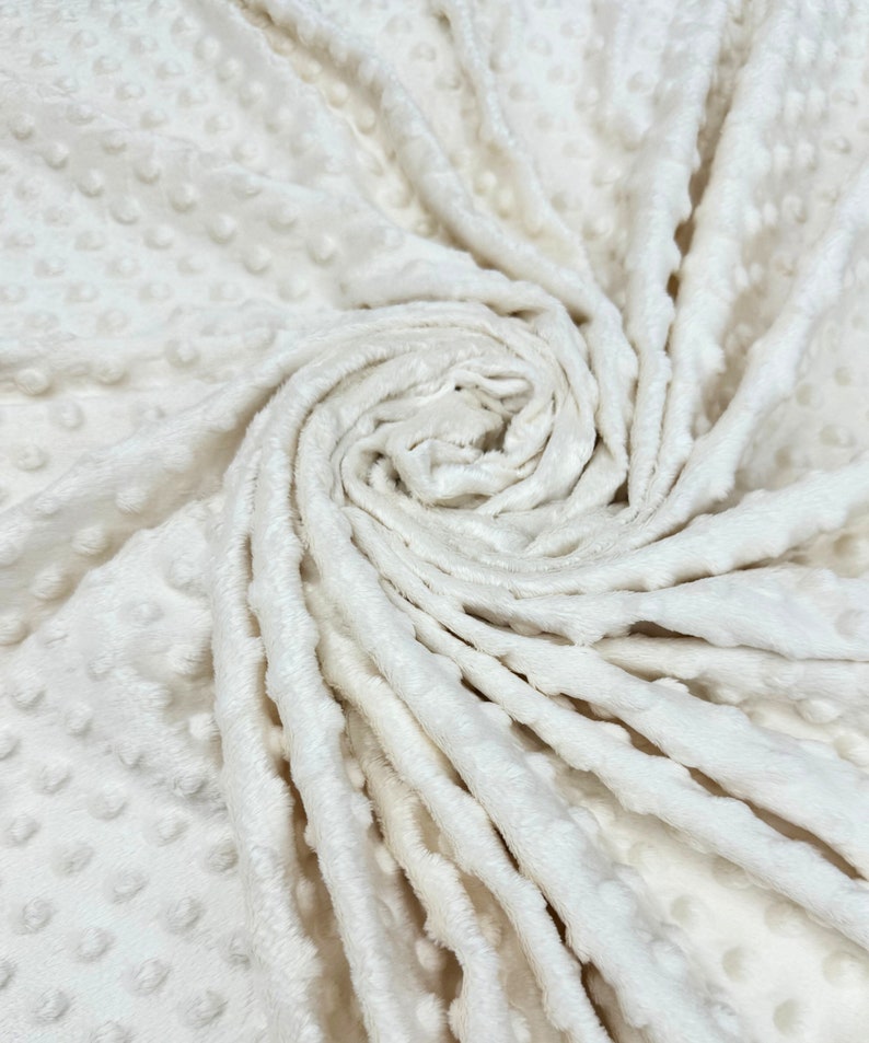 May include: Close-up of a white, soft, textured fabric with a raised dot pattern. The fabric is folded and twisted, showing the texture and softness.