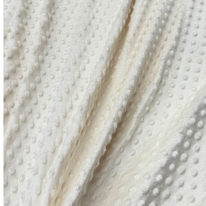 May include: Close-up of a white, textured fabric with a raised dot pattern. The fabric is soft and plush, and it has a subtle sheen.