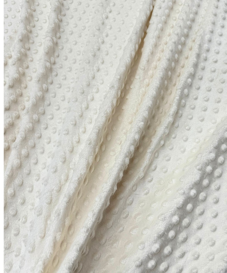 May include: Close-up of a white, textured fabric with a raised dot pattern. The fabric is soft and plush, and it has a subtle sheen.