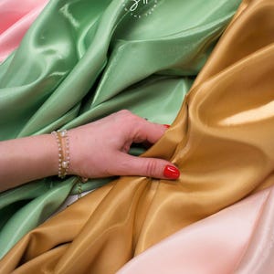 May include: A hand with red nail polish rests on folds of shimmering fabric in shades of gold, green, and pink. A delicate gold bracelet with pearls adorns the wrist. The fabric appears to be silk or satin.