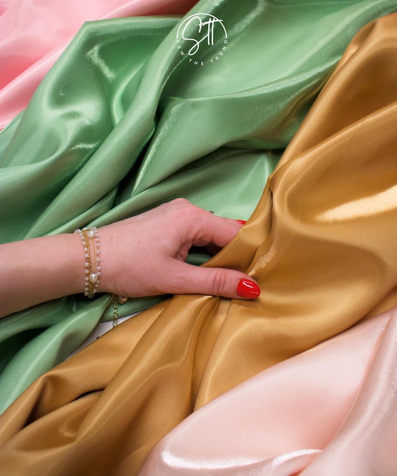 May include: A hand with red nail polish rests on folds of shimmering fabric in shades of gold, green, and pink. A delicate gold bracelet with pearls adorns the wrist. The fabric appears to be silk or satin.