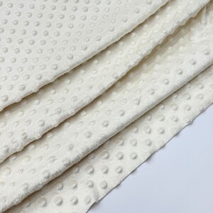 May include: Close-up of a white, soft, textured fabric with a subtle dot pattern. The fabric is folded in layers, showcasing its plush texture.