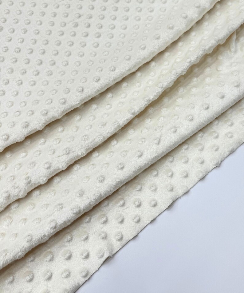 May include: Close-up of a white, soft, textured fabric with a subtle dot pattern. The fabric is folded in layers, showcasing its plush texture.